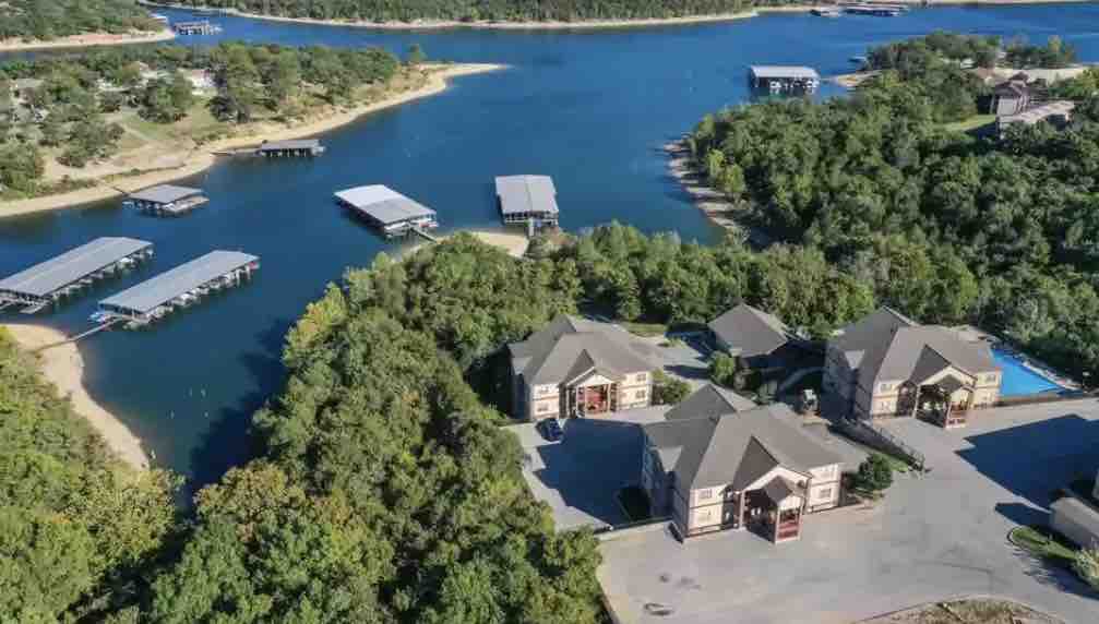 An aerial view captures the surrounding natural landscape with Table Rock Lake and several boat docks nestled among lush greenery. The property layout includes two distinct condo buildings, complemented by a swimming pool and easy access to the water.