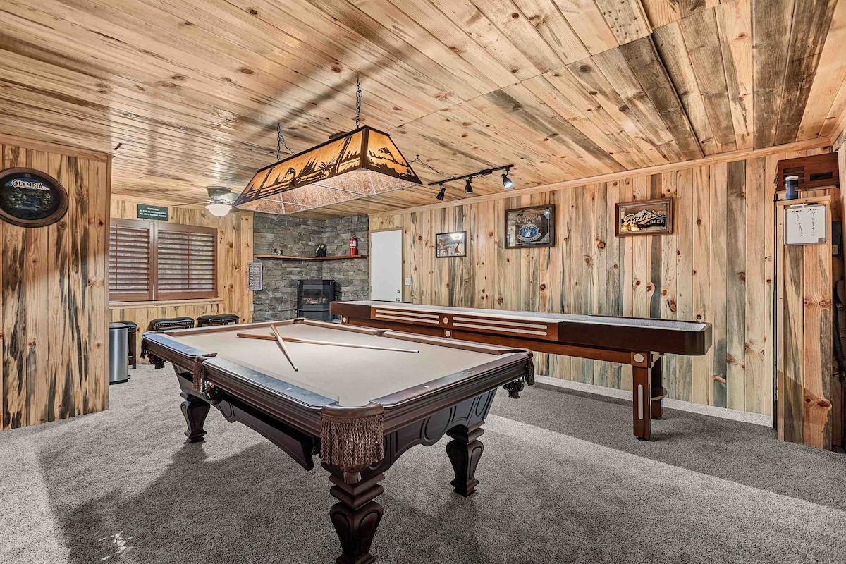 The game room features a billiards table and a shuffleboard table, both positioned on plush carpet. Textured wood paneling adorns the walls, complemented by soft lighting from overhead fixtures. A stone fireplace adds an element of warmth, enhancing the inviting setting for leisure activities.