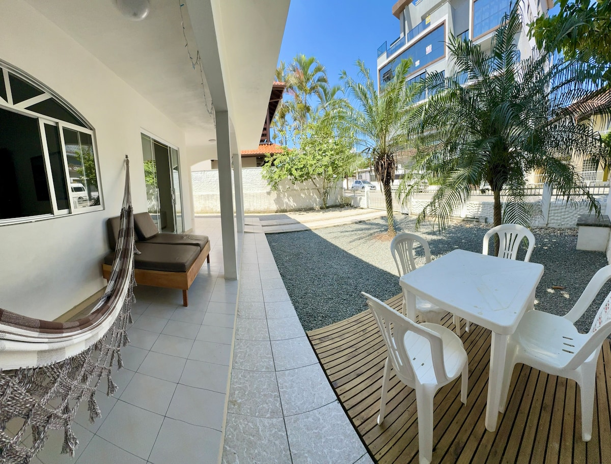 An outdoor area features a wooden deck with a white table and four chairs, surrounded by gravel and palm trees. A spacious bed and a hammock are positioned on the covered patio, providing a relaxed space for leisure. Bright sunlight enhances the welcoming atmosphere.