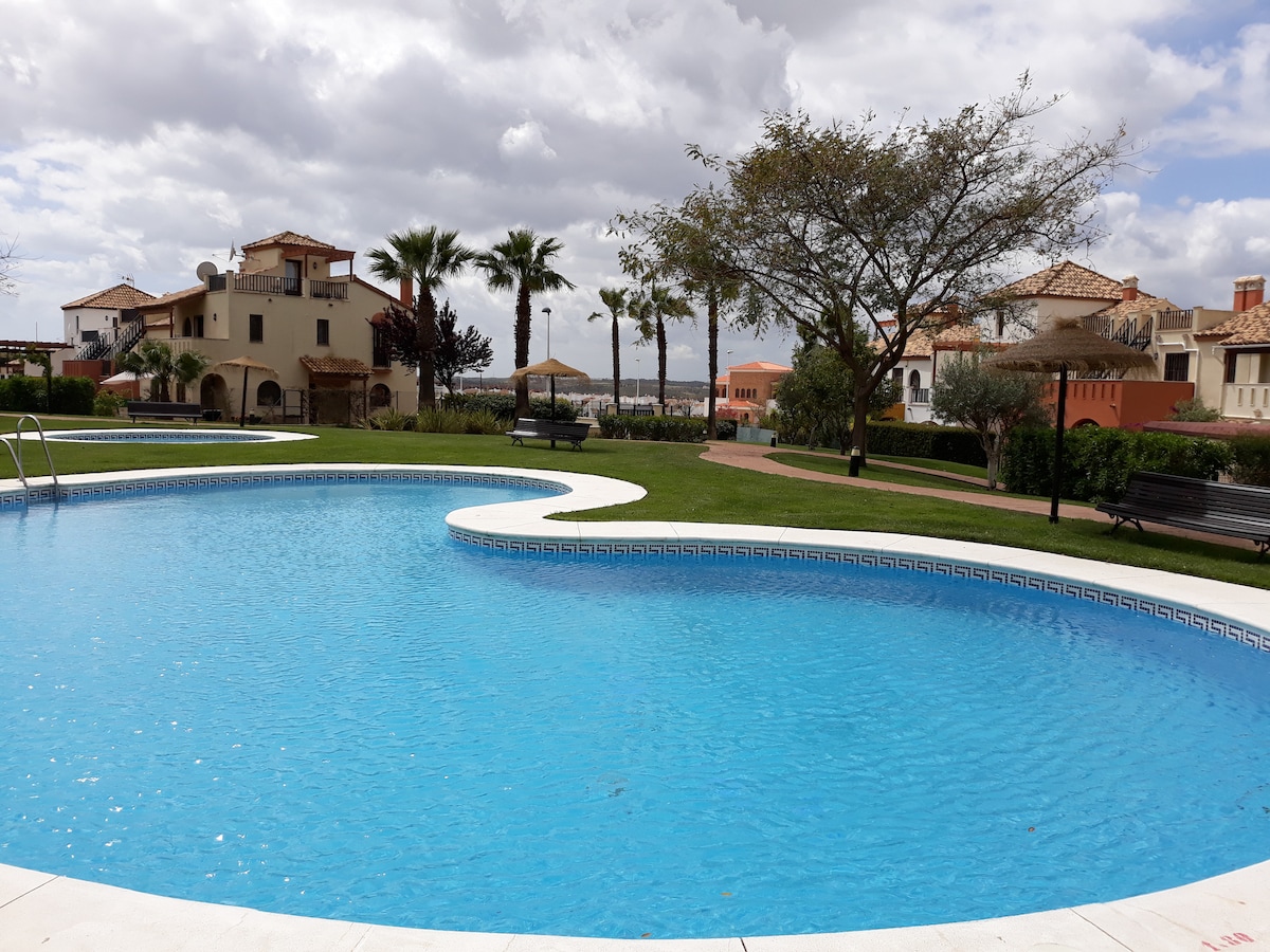 The community pool features a curved shoreline with clear blue water, surrounded by well-maintained green grass and palm trees. Lounge chairs are positioned along the poolside, offering space for relaxation. The background showcases residential buildings with terracotta roofs and landscaping, contributing to a serene atmosphere.