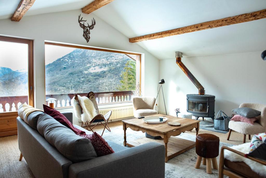 The living room features large windows offering views of the surrounding mountains. A comfortable sofa and various seating options are arranged around a wooden coffee table. A wood-burning stove adds warmth, while the ceiling showcases wooden beams for a rustic touch.
