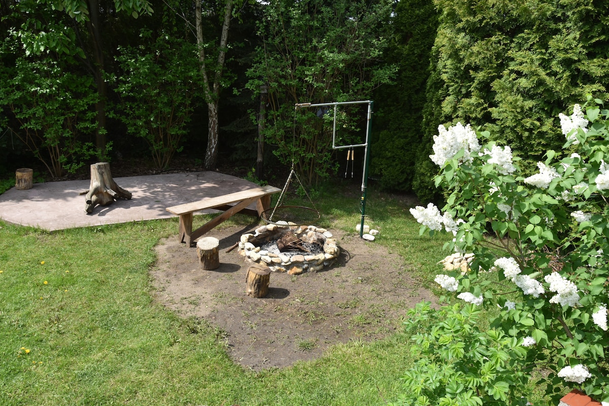 A peaceful garden features a wooden fire pit surrounded by stones and seated on natural stumps. A small wooden platform nearby offers additional gathering space, while lush greenery and flowering plants enhance the serene outdoor setting.