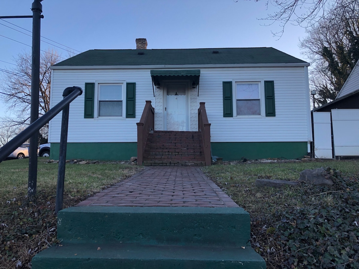 A single-story home is presented with a green and white exterior, featuring a front porch with wooden stairs leading to a central double door. Flanking the entrance are two large windows with green shutters, and a well-maintained pathway made of paving stones is visible.