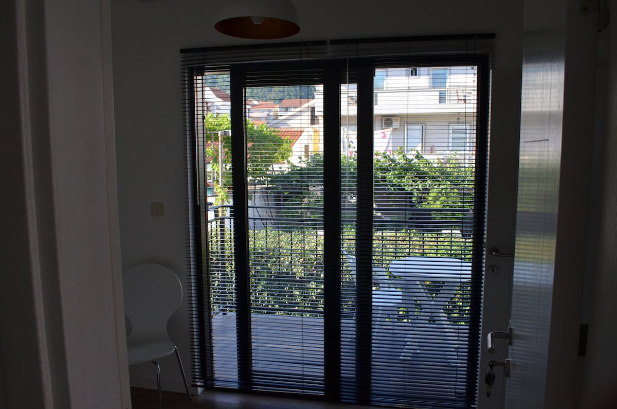 A set of large glass doors framed by black blinds opens onto a small balcony. A white table and a chair are visible on the balcony, surrounded by greenery from plants outside.