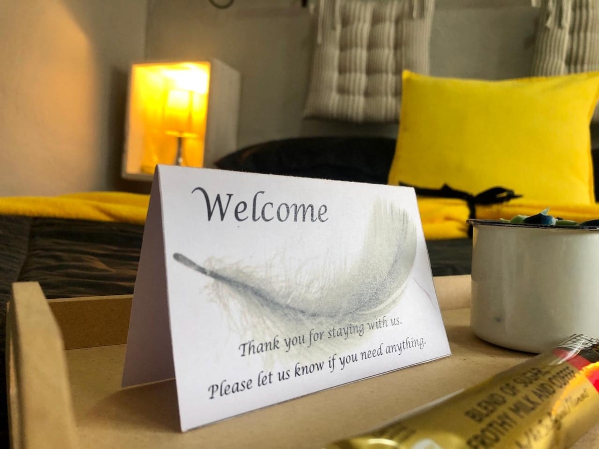 A welcome card is positioned on a tray, featuring a simple feather design and an inviting message. A small candle illuminates the background, with a glimpse of yellow and neutral tones from the bedding and wall decor, creating a calm and cozy ambiance.