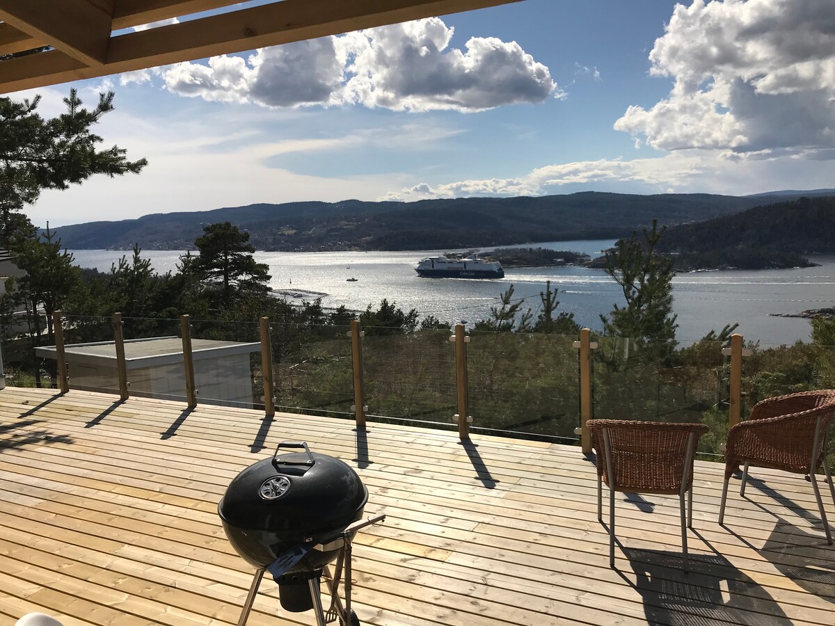 A spacious wooden terrace features a coal grill and two woven chairs, offering stunning views of the fjord and surrounding landscape. The area is sunlit, with large glass railings ensuring unobstructed vistas of the water and distant hills.