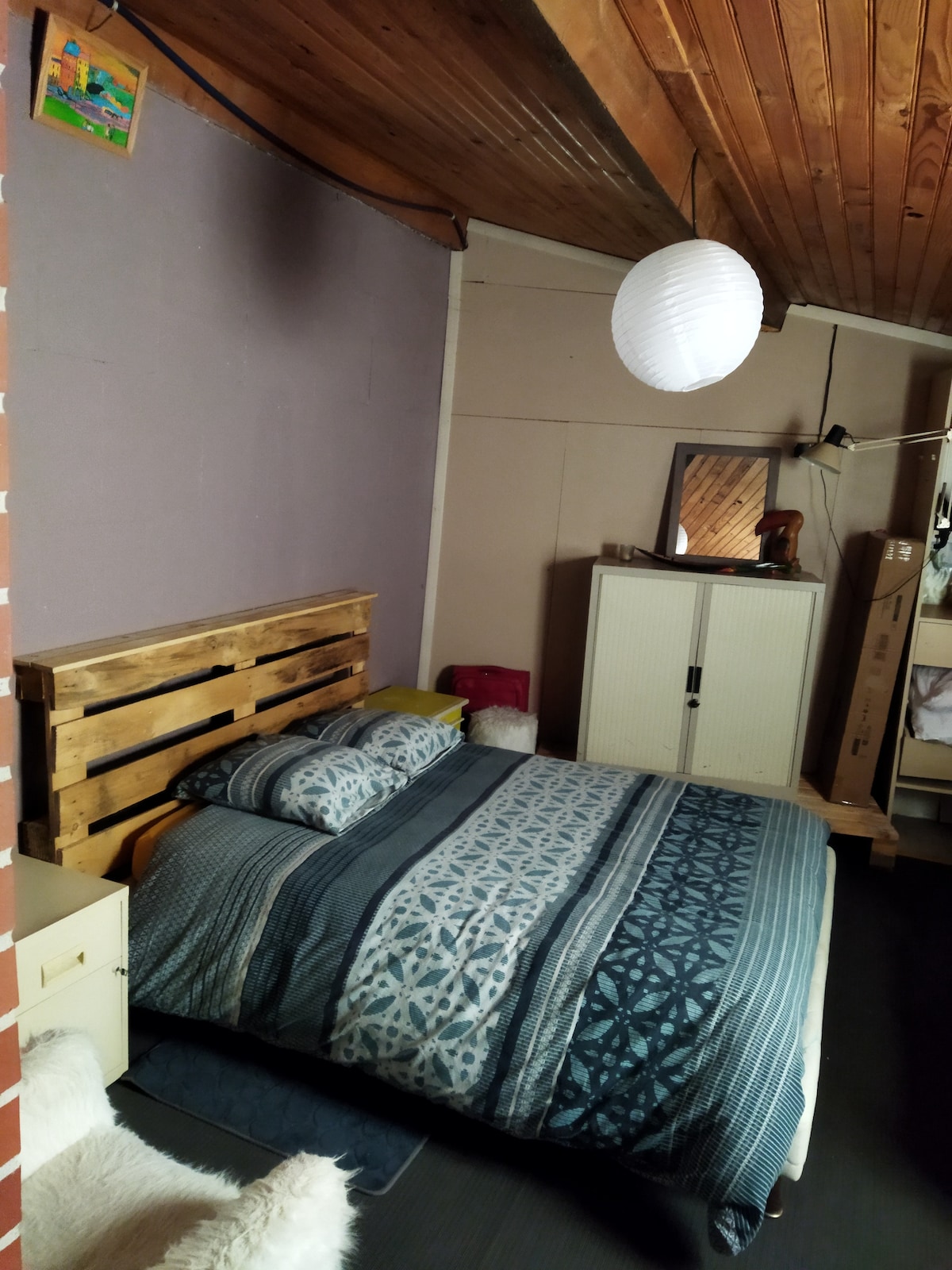 The cozy sleeping area is characterized by a wooden pallet bed adorned with patterned bedding and two pillows. A simple mirror is mounted on the wall above a white cabinet, while a paper lantern hangs from the ceiling, contributing to the room's warm ambiance.