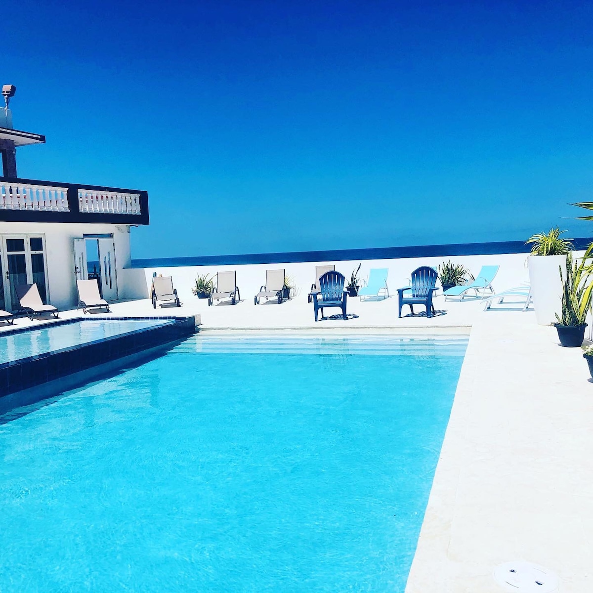 A small private pool is situated beside a larger pool, surrounded by a patio with sun loungers. The expansive sky stretches overhead, showcasing a clear blue hue. Soft seating can be noted on the balcony area adjacent to the pools.