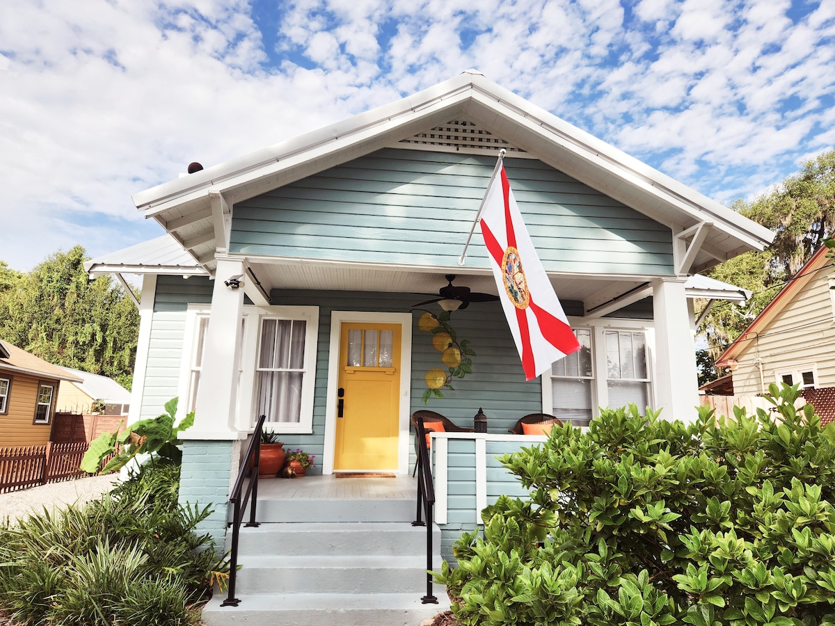 The exterior of the bungalow features a pale blue façade with white trim and a welcoming yellow door. A Florida flag hangs prominently, and two plants sit on the porch steps. Steps with handrails lead to the spacious front porch, surrounded by lush greenery.