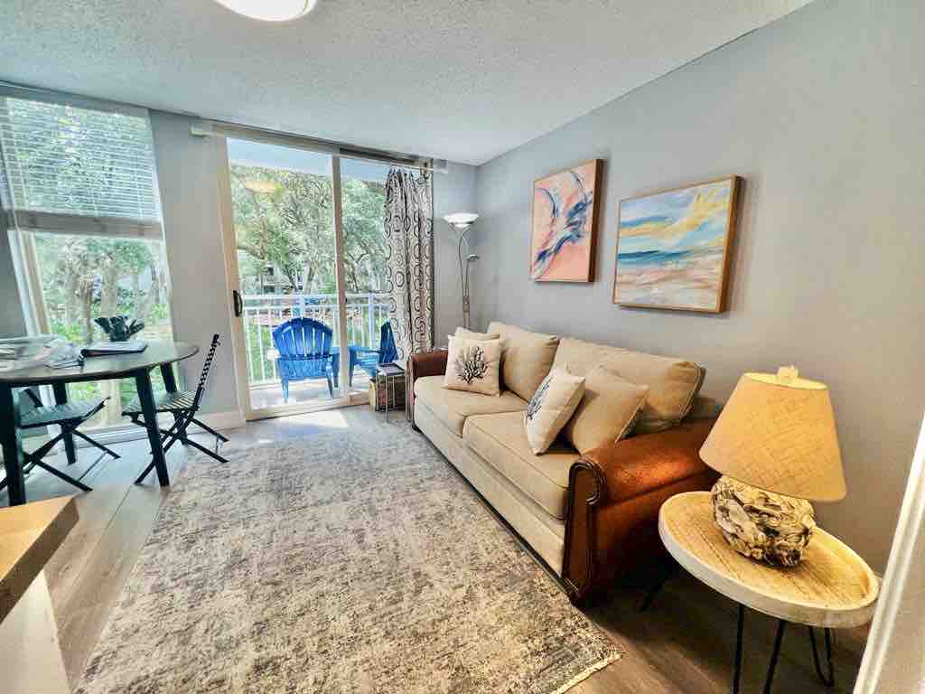 A bright living area features a comfortable sofa accompanied by a side table with a lamp. Colorful artwork adorns the walls, and natural light filters in through sliding glass doors that open to a balcony. A small dining table with chairs is visible in the background.