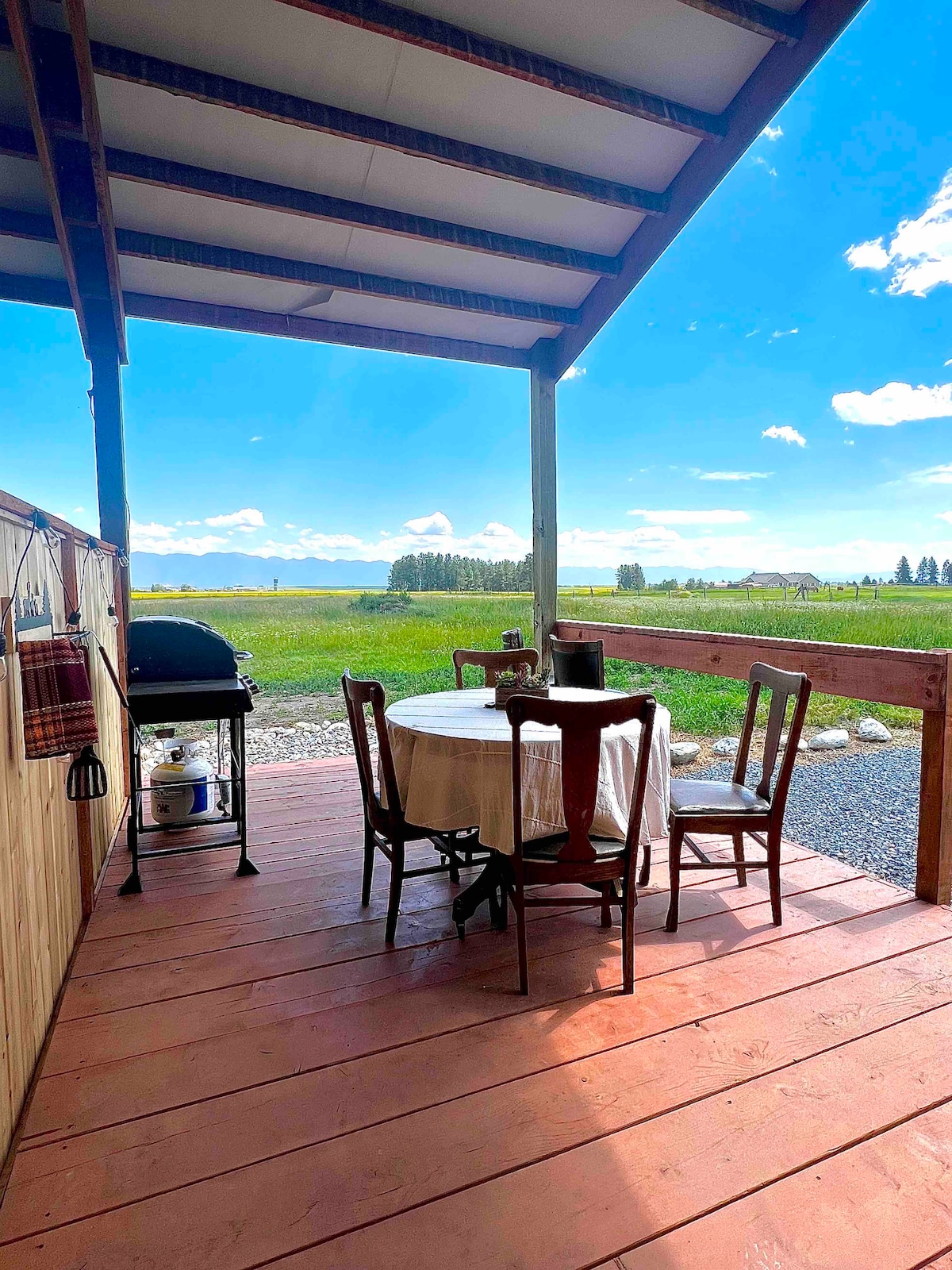 A covered patio area features a round table surrounded by four wooden chairs. Nearby, a grill is positioned on a stone surface. A scenic landscape of green fields and distant trees is visible in the background under a clear blue sky.