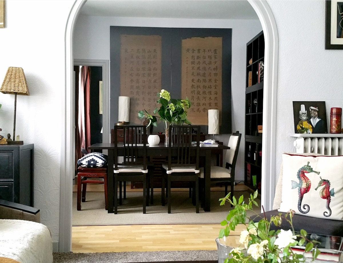A modern dining area is visible through an archway, featuring a dark wooden table surrounded by six chairs. On the table, a decorative vase holds fresh flowers. The walls are adorned with scroll-like artwork, adding a touch of elegance to the space.
