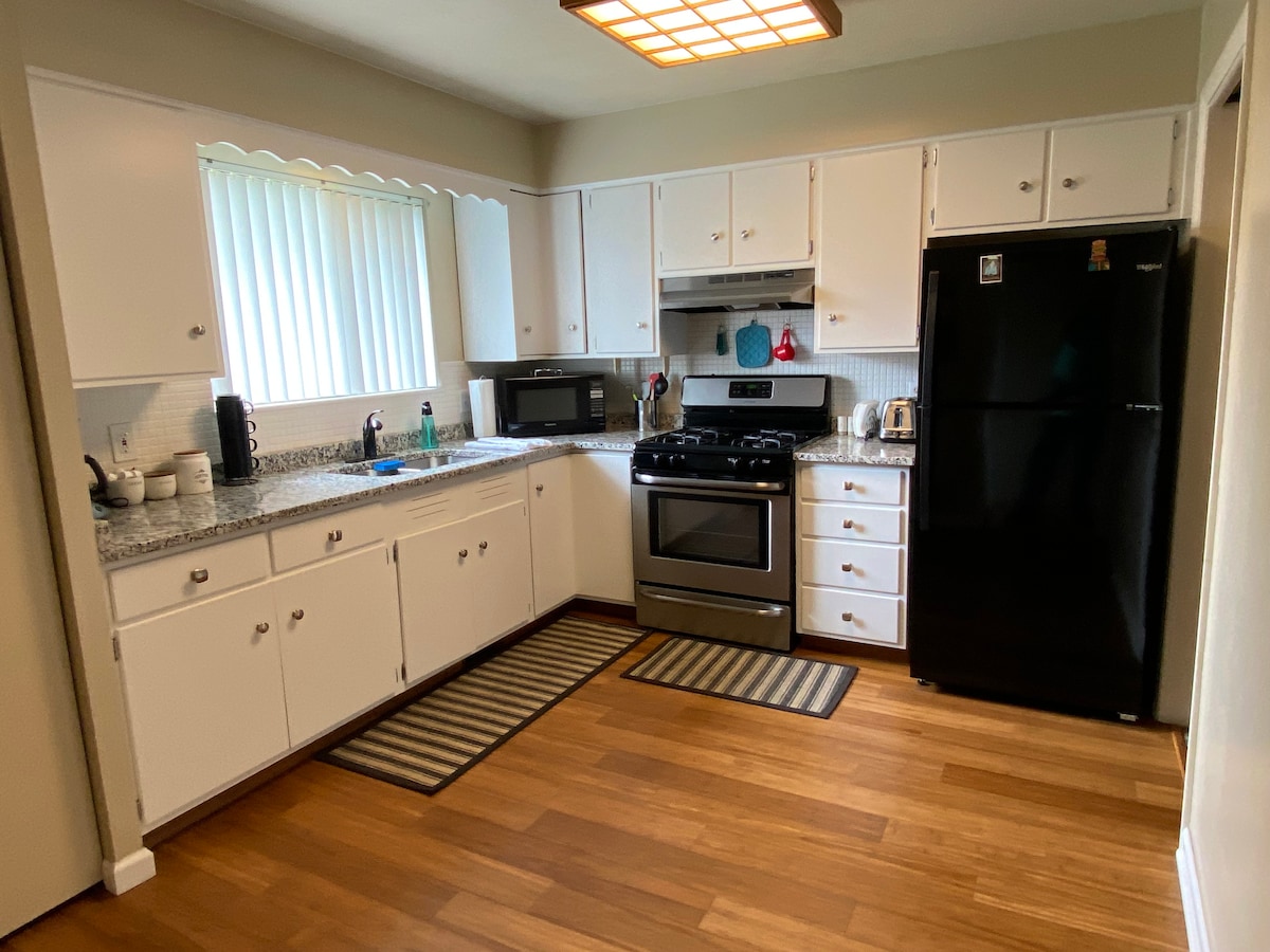The kitchen is equipped with white cabinetry and modern appliances. A black refrigerator and a gas stove are featured, along with a microwave and countertop space. Natural light enters through a window adorned with vertical blinds, while wooden flooring and area rugs add warmth to the space.