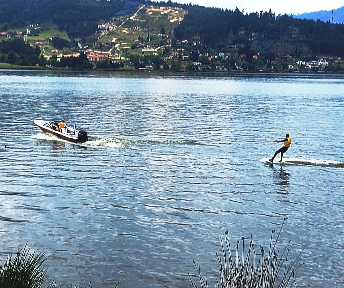 A person is seen water skiing behind a motorboat on the calm surface of a lake. The background features lush green hills and distant houses, with the sky partially covered by clouds. Grasses line the water's edge, adding to the natural landscape.