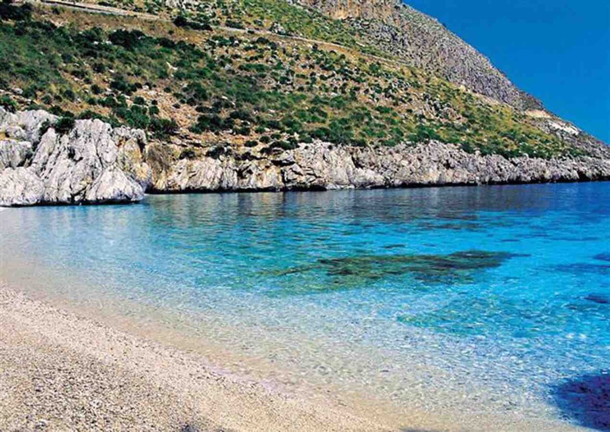 Calm waters are viewed along a rocky coastline, with gentle waves lapping at the sandy shore. The clear blue hues of the sea complement the vibrant greenery of the hillside in the background, creating a serene and natural setting.