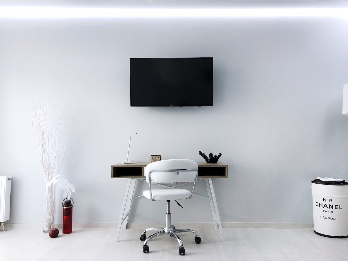A work area is presented with a modern desk and a comfortable office chair. Above, a mounted flat-screen TV is visible. Minimalist decor includes a white display vase and a stylish waste bin, enhancing the overall contemporary design of the space.