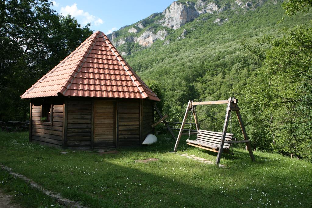 A rustic wooden structure with a distinct triangular roof is nestled among lush greenery. A swing set is positioned nearby on the grassy area, with a mountainous landscape visible in the background under a clear blue sky.