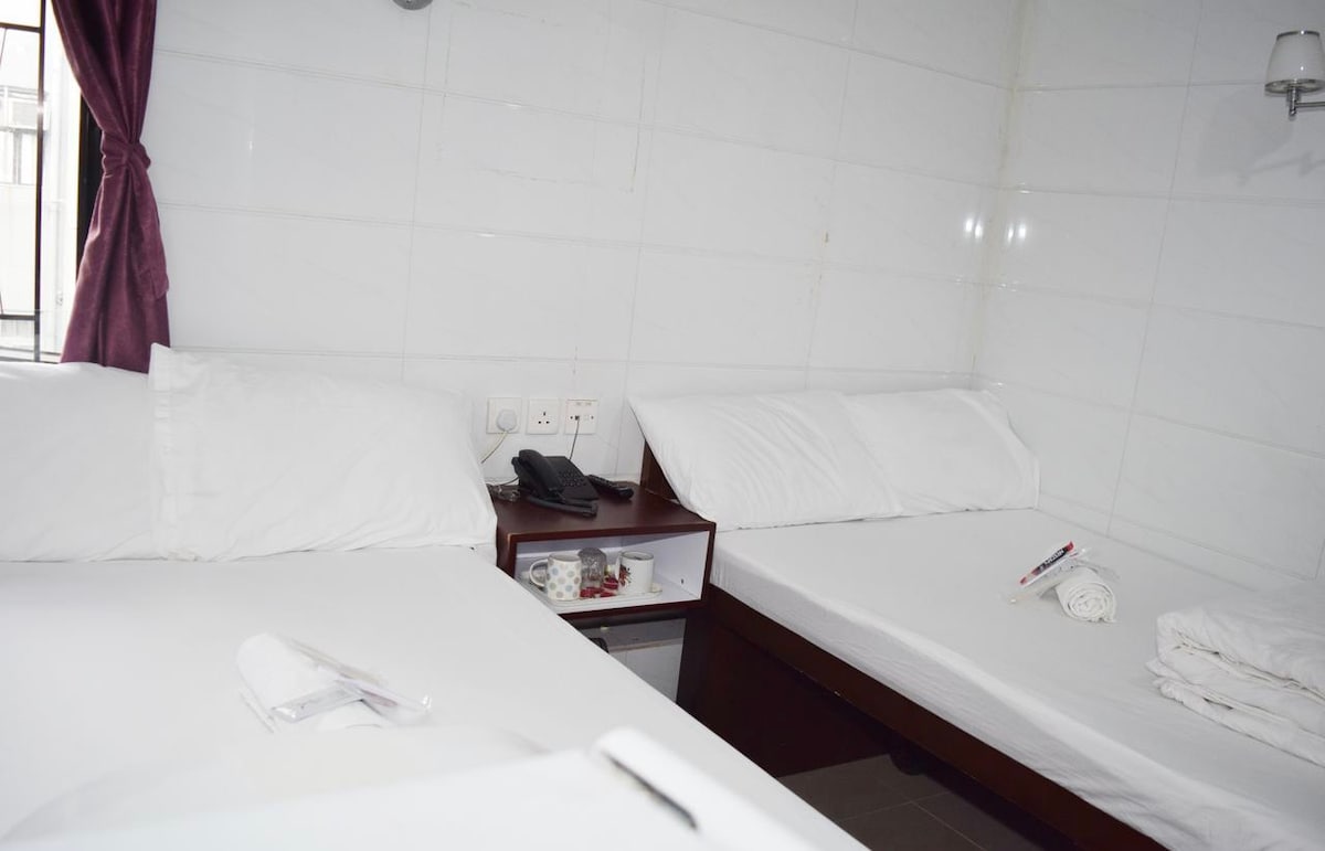 The room features two spacious beds arranged side by side, with clean white linens. A nightstand is positioned between the beds, containing a telephone and small storage. Natural light enters through a nearby window covered by a deep red curtain.