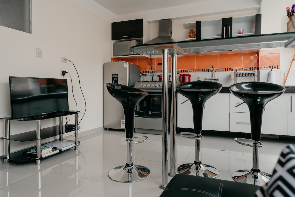 A modern kitchen is shown, featuring sleek black bar stools around a glass-top counter. An array of kitchen appliances and utensils is visible, alongside a television on a stand. The clean, bright space is enhanced by orange accents on the wall.