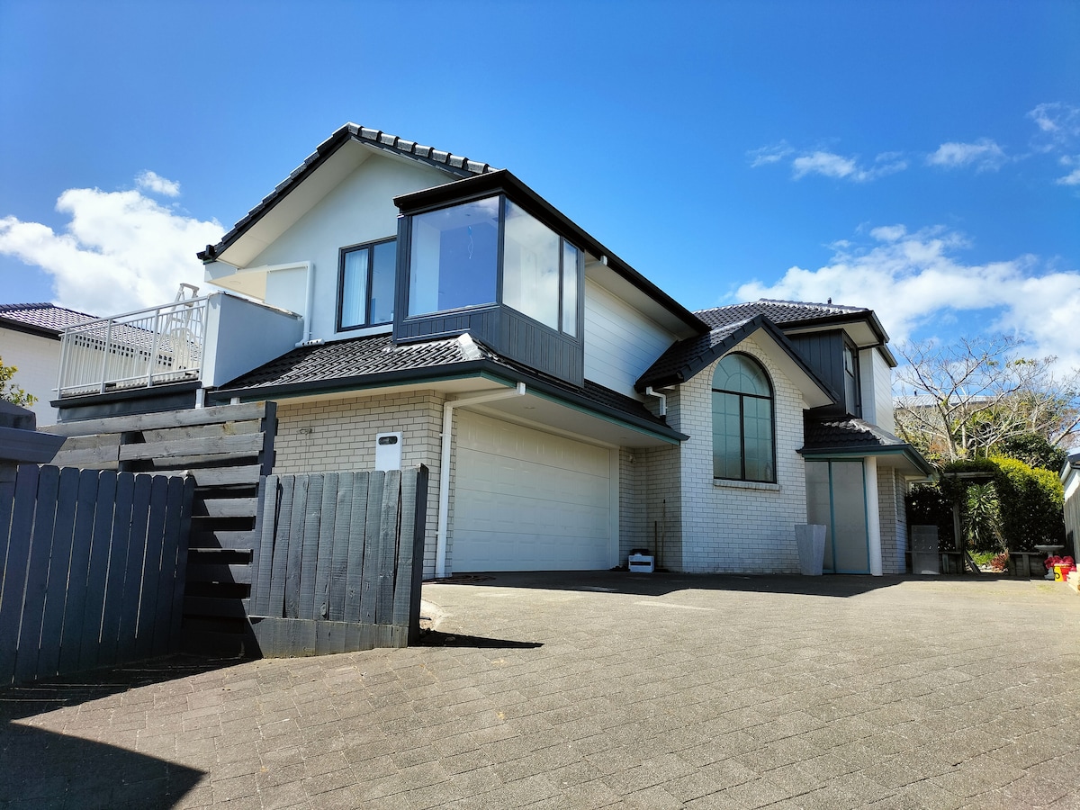 An exterior view of a spacious two-story home is displayed, featuring a driveway and garage. A combination of white brick and dark roofing materials is visible. Large windows allow natural light, and a secluded outdoor area is suggested by the surrounding greenery.