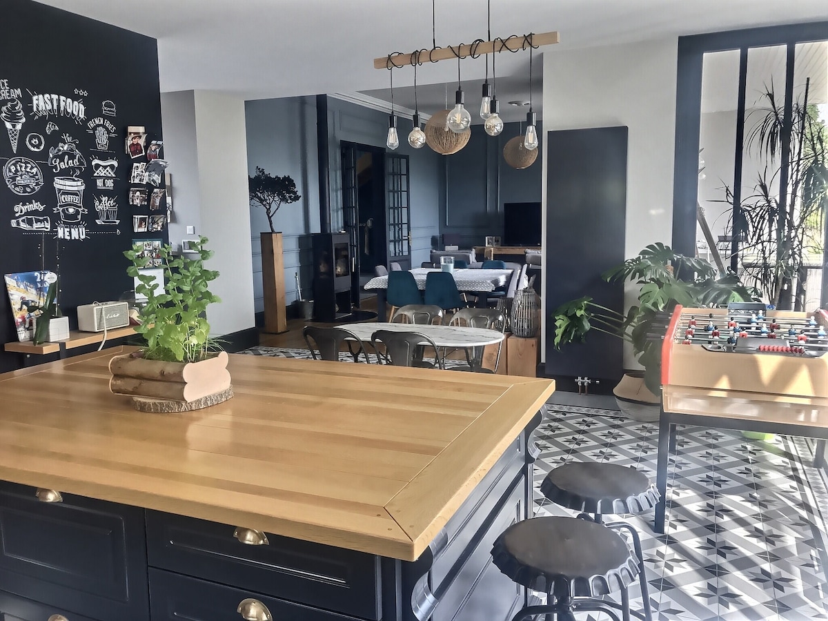 A spacious kitchen is featured with a wooden island and bar stools. Black cabinetry and wall accents add contrast to the open layout. The adjoining dining and living areas are visible, characterized by modern furnishings and decorative plants, with a foosball table positioned in the corner.
