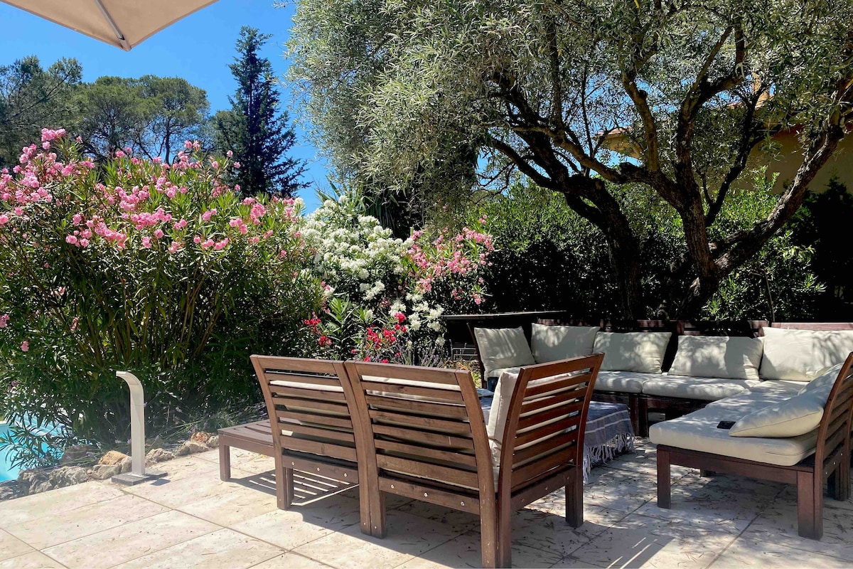An outdoor seating area features comfortable lounge sofas arranged around a coffee table, surrounded by lush greenery and colorful flowering plants. The setting is shaded by an umbrella while a glimpse of the turquoise pool is visible in the background.