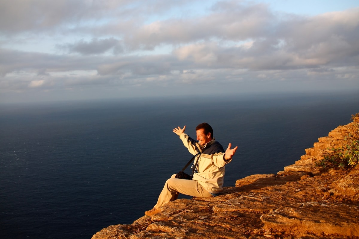 A person is seated on a rocky ledge overlooking a vast ocean, with arms spread wide. The calm sea reflects soft sunlight and the sky is partially cloudy, displaying various shades of blue and gray. The surrounding landscape features rugged rocks and sparse vegetation.