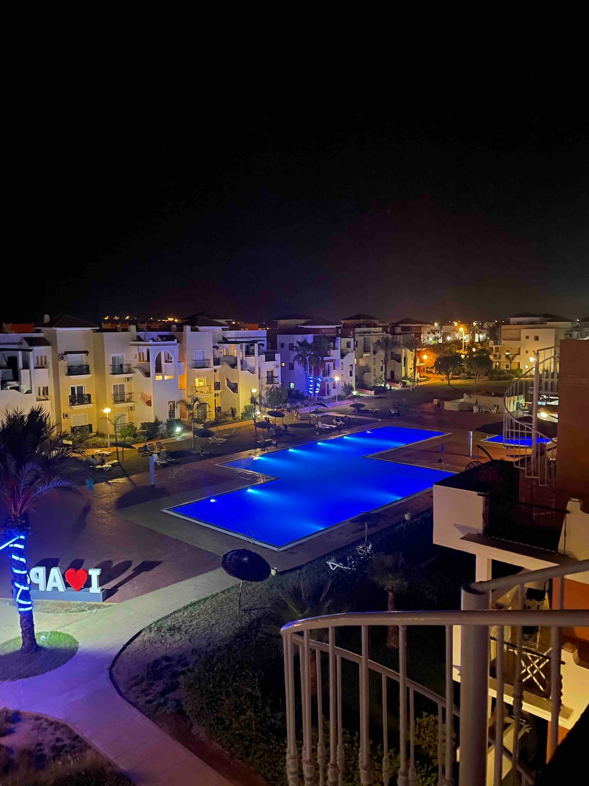 A night view captures a private residence with illuminated buildings surrounding a spacious pool glowing in blue. Decorative lights add a soft ambiance to the area, while palm trees frame the scene, enhancing the tranquil environment.