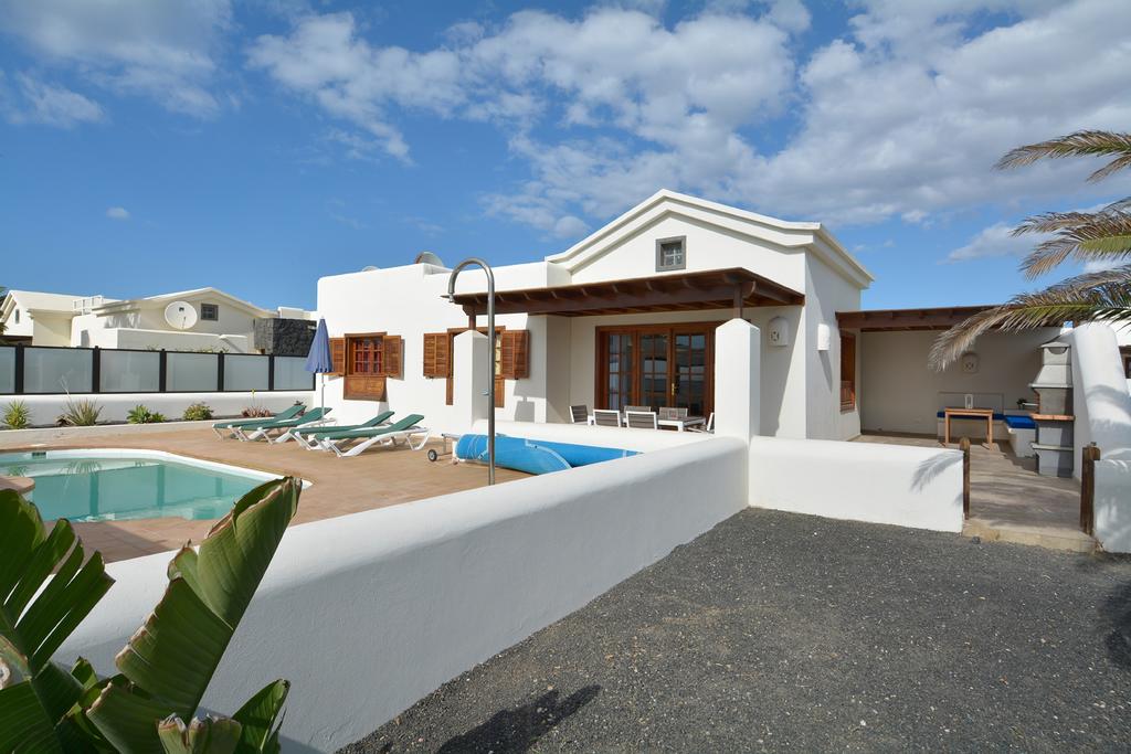 An exterior view of a white villa with a terracotta roof, surrounded by a wooden deck and a tranquil pool. Sun loungers are arranged by the water, with palm fronds framing the foreground. The sky is clear with scattered clouds, enhancing the peaceful setting.