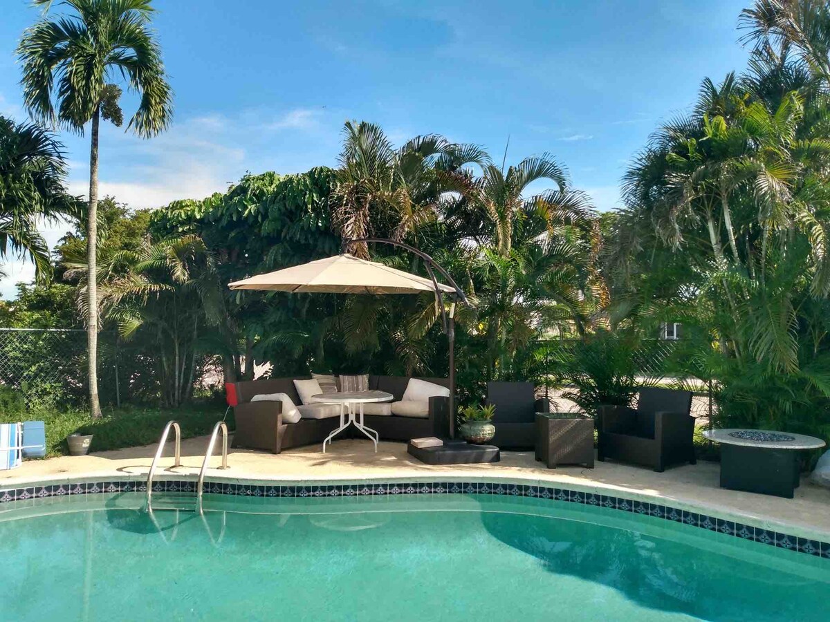 A serene pool area is surrounded by lush tropical greenery. A sandy-colored patio features a shaded seating arrangement under an umbrella, accompanied by comfortable outdoor chairs. The pool water reflects the bright sky, inviting relaxation in a tranquil setting.