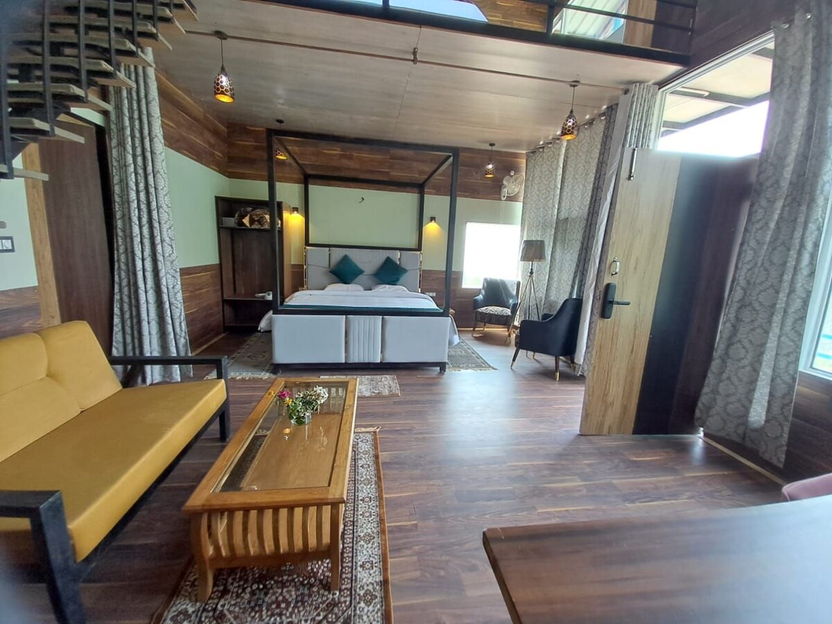 A spacious room features a canopy bed with plush pillows and light bedding. A cozy seating area includes a yellow sofa and wooden coffee table. Large windows allow natural light to fill the space, complemented by warm wood accents and decorative curtains.