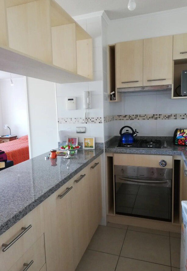 The kitchen features light wood cabinetry and a granite countertop. Under-cabinet lighting illuminates the preparation area. A kettle sits on the stove, and various appliances are neatly arranged. Mosaic tiles provide a decorative touch above the countertop, enhancing the practical layout.