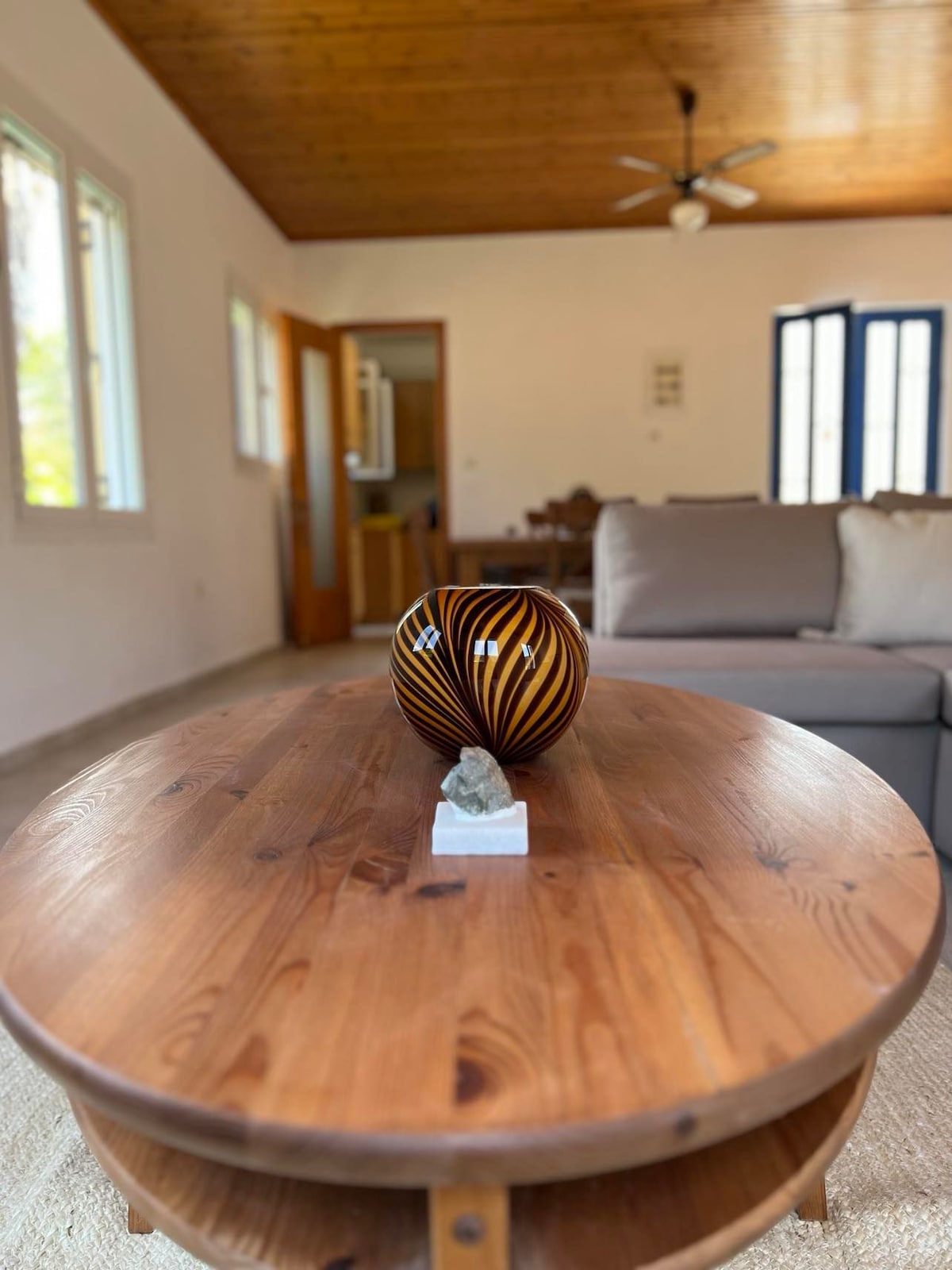 A wooden coffee table is centered in the living space, featuring a striped decorative vase and a small sculptural piece. The room is brightened by natural light from multiple windows, while comfortable seating is arranged nearby, creating an inviting area for relaxation.