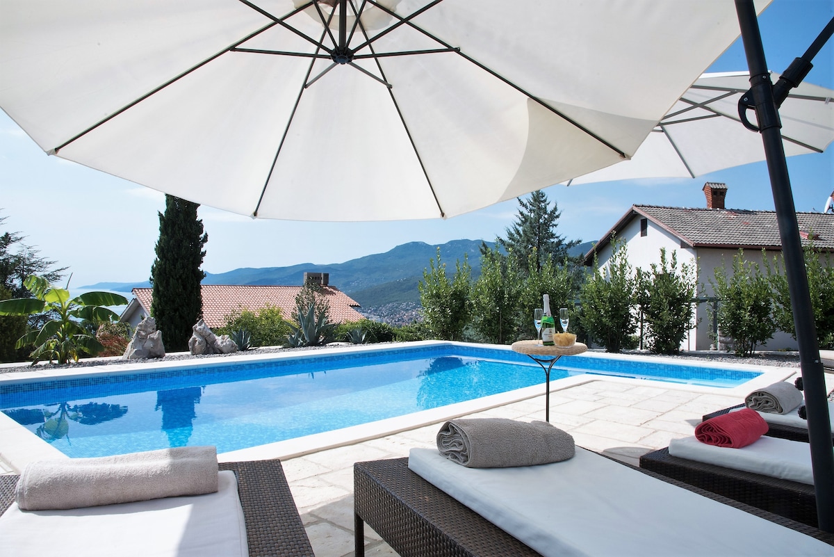 A serene pool area is highlighted under large umbrellas, offering shade for loungers. A calming blue pool reflects the clear sky, surrounded by lush greenery and distant hills. Soft towels are neatly arranged on the loungers, providing a welcoming space for relaxation.