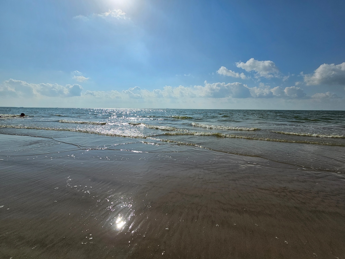 An expansive view of a serene beach scene under a clear blue sky. Soft waves gently lap against the shore, and sunlight sparkles across the water's surface, creating a shimmering effect. A distant figure is visible, enjoying the tranquil atmosphere.