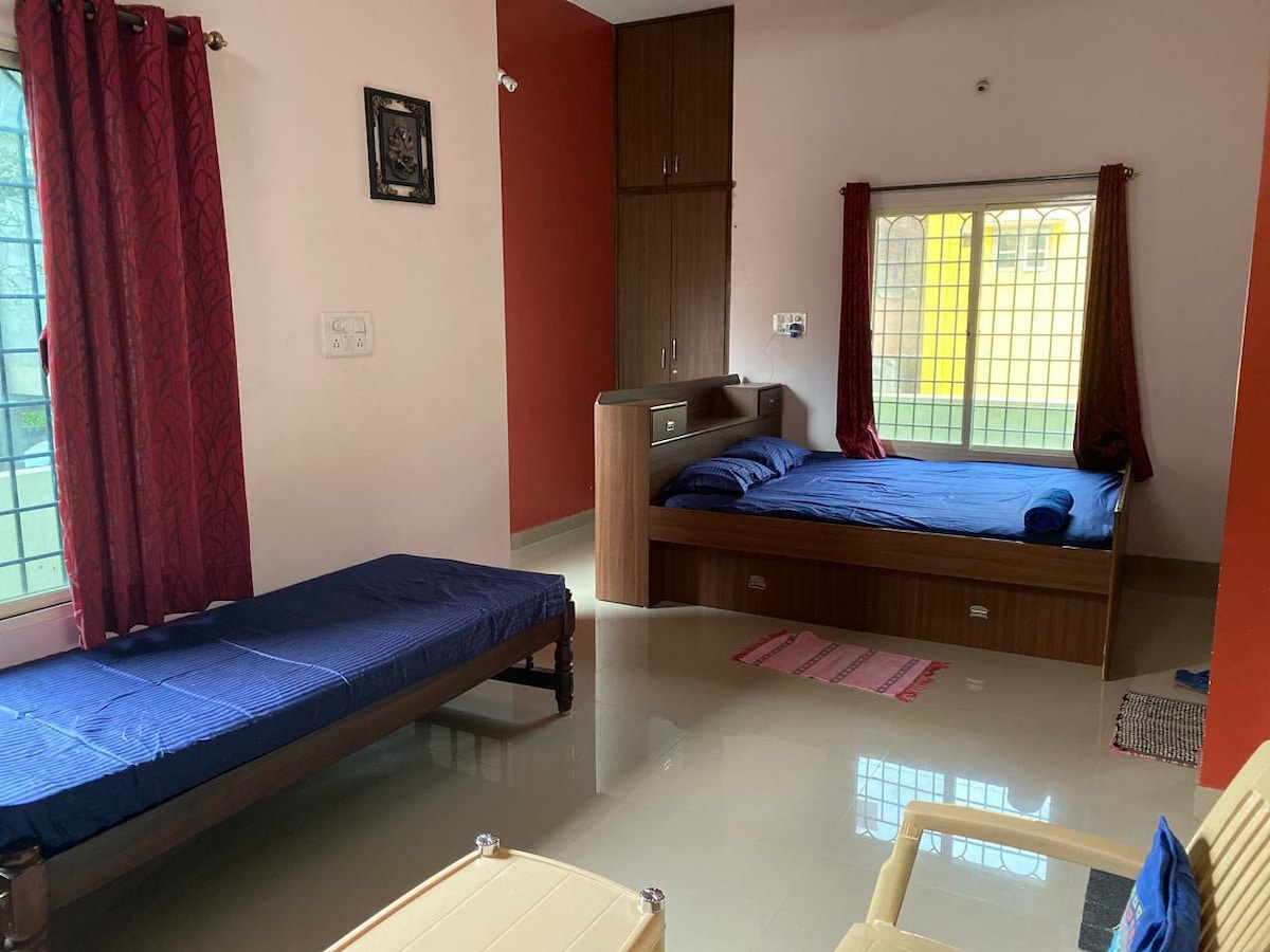 The room features a spacious layout with one large bed and an additional smaller bed, both covered in blue linens. Natural light fills the space, highlighting the well-maintained floor and colorful curtains. A small side table is positioned against the wall, enhancing functionality.