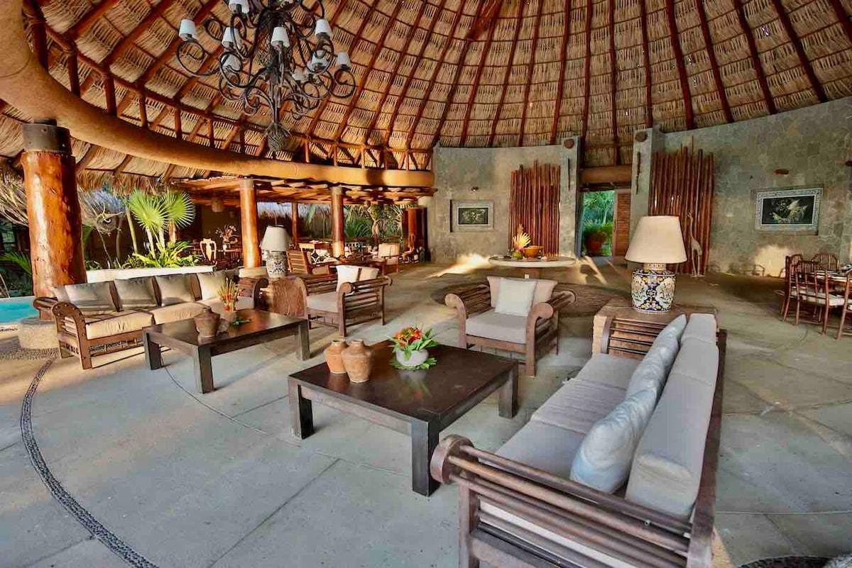 A spacious outdoor living area is featured with high thatched ceilings and natural wooden beams. Comfortable seating arrangements are provided, with decorative lamps placed on side tables. Potted plants and tropical greenery surround the space, enhancing the connection to nature.