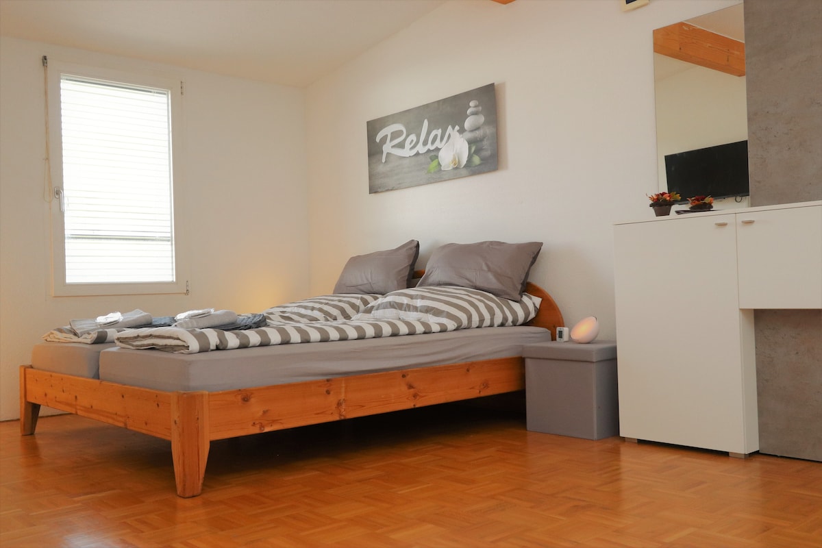 A spacious sleeping area is featured with a wooden bed frame and striped bedding. A wall-mounted artwork with the word 'Relax' enhances the room. Natural light enters through two windows, while a small nightstand and a storage unit are present, contributing to the room's functionality.