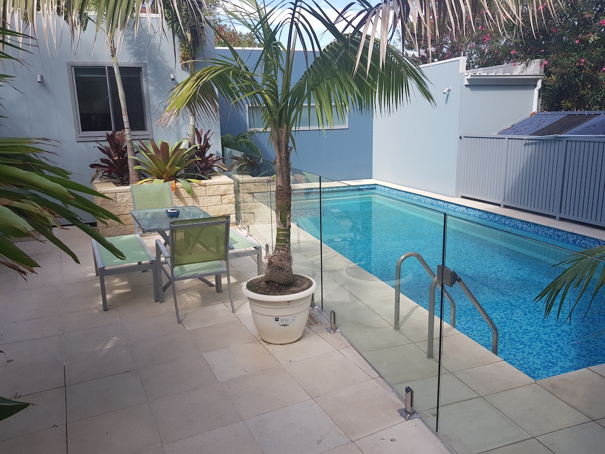 An outdoor area features a large swimming pool surrounded by a glass railing. Comfortable lounge chairs are positioned beside the pool, while tropical plants enhance the setting. Natural light reflects off the water, creating a relaxing atmosphere.