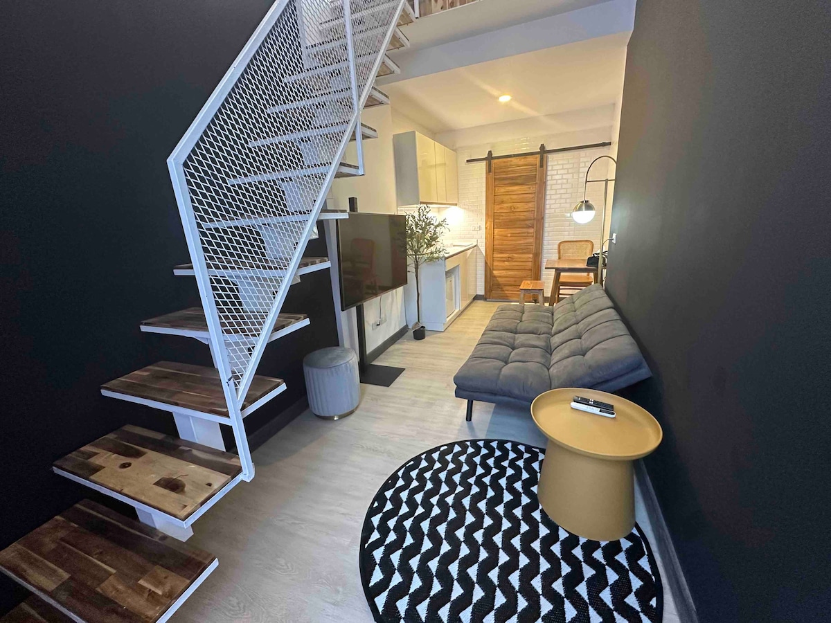 A modern living area is showcased, featuring a comfortable sofa and a unique round side table. Open shelving and a staircase lead to the upper level, while a sleek kitchen area is visible in the background. A patterned rug adds texture to the space.