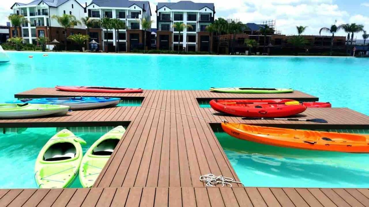 A wooden floating deck is positioned on a tranquil lagoon, with brightly colored kayaks resting beside it. The clear blue water surrounds the deck, enhancing the inviting atmosphere for outdoor activities. Lush greenery and modern buildings are visible in the background.