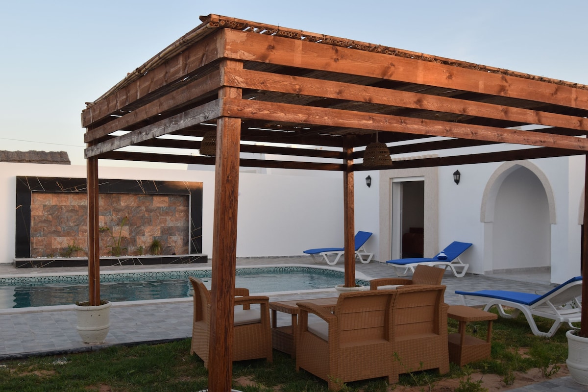 A shaded terrace features a wooden pergola supported by sturdy posts, surrounded by seating and sun loungers. The tranquil pool area is visible in the background, complemented by a stone feature wall that adds a touch of elegance.