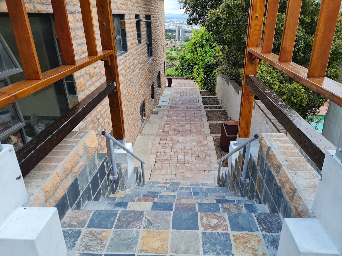 A stone pathway lined with greenery descends from a wooden deck, leading to a paved walkway. The stairs are flanked by sturdy wooden railings. View of the surrounding hillside and distant city skyline is visible in the background.
