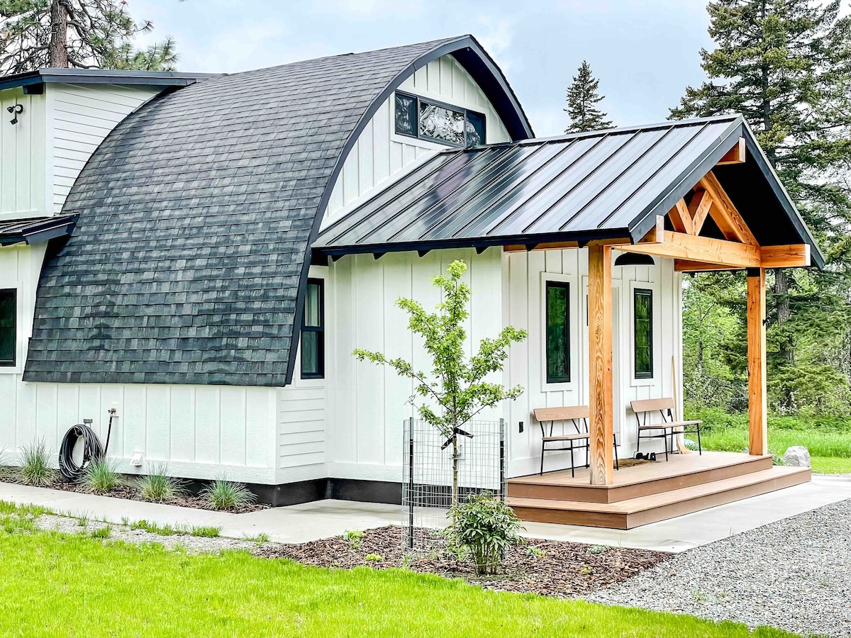 The cottage exterior features unique whalebone architectural design with a curved roof and a welcoming front porch. Two benches provide seating, set against a backdrop of green grass and framed by trees, highlighting the peaceful surroundings.