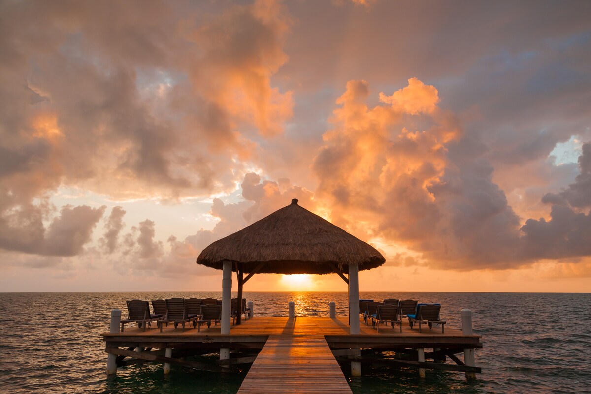 A spacious wooden pier extends over the water, featuring a thatched-roof gazebo at its end. Several lounge chairs are arranged neatly, facing the horizon where warm tones of sunset illuminate the sky, blending shades of orange and pink with soft clouds.