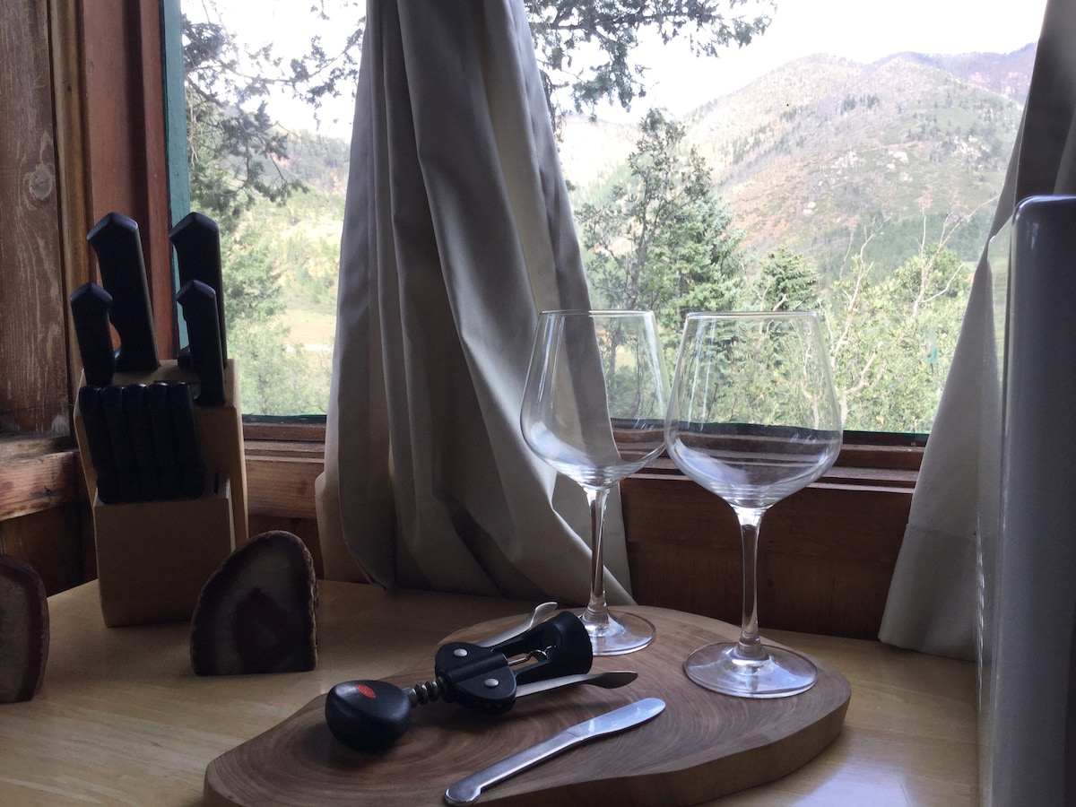 Two glass wine glasses are placed on a wooden cutting board next to a corkscrew and knife, with a backdrop of mountains visible through the window. Knives are stored in a wooden block nearby, complemented by light curtains gently framing the view.