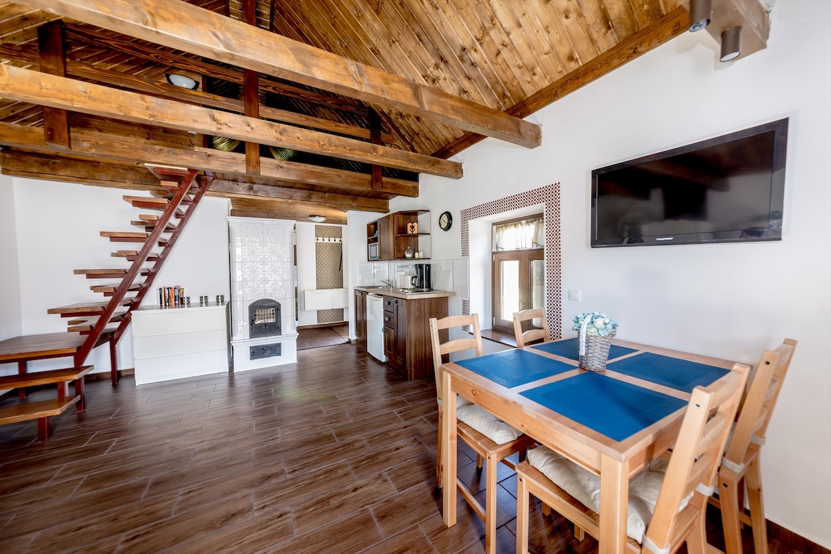 An open-plan living area features wooden beams and a high ceiling. A dining table with blue placemats is positioned near a kitchen area, which includes modern appliances. A staircase leads to an upper level, while a fireplace adds warmth to the space. A television is mounted on the wall.