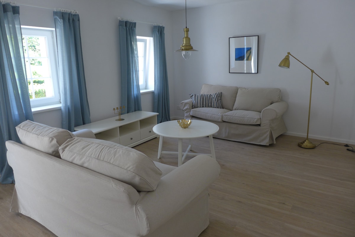 A bright living room features two light-colored sofas positioned around a round coffee table. Large windows dressed in blue curtains allow natural light to fill the space. A minimalist light fixture hangs above, and a piece of wall art adds a subtle touch of decor.