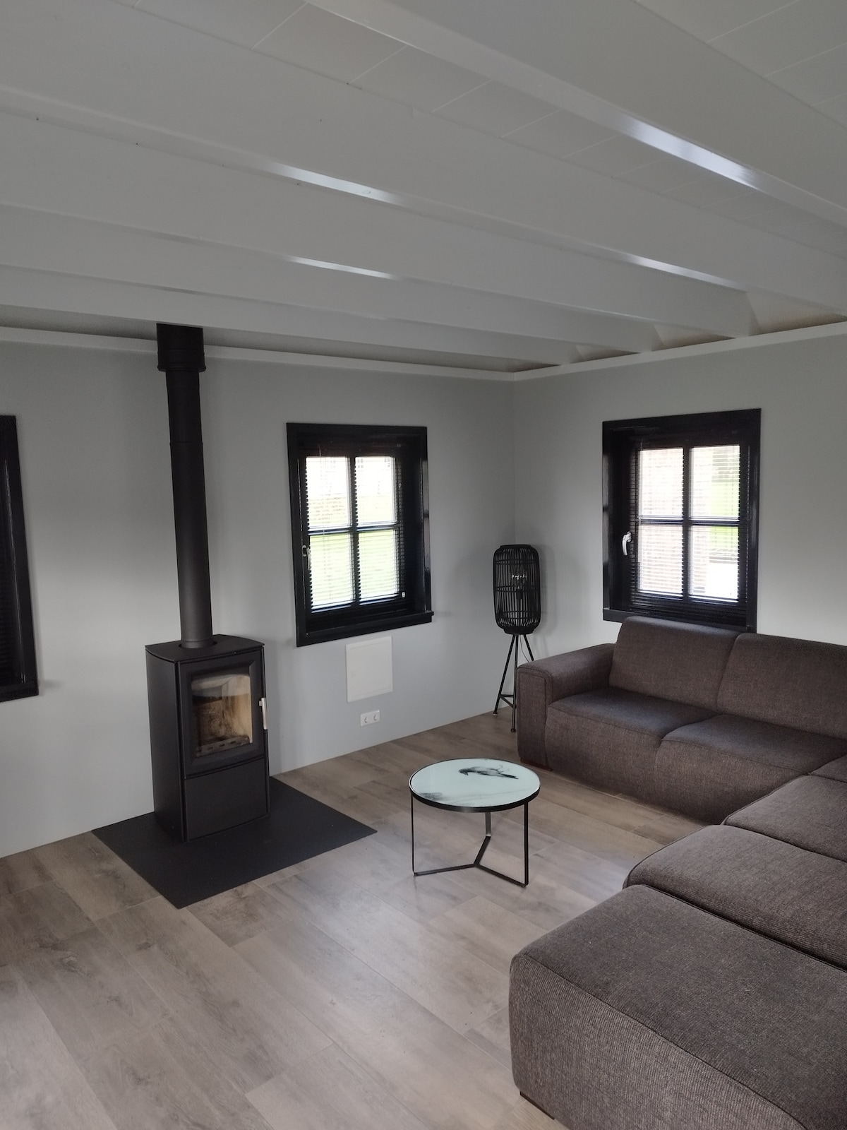 A modern living area features a dark sofa arranged around a small circular coffee table. A sleek wood-burning stove stands on a dark base, complemented by two black windows that allow natural light to filter in. Light-colored wooden flooring enhances the spacious feel.