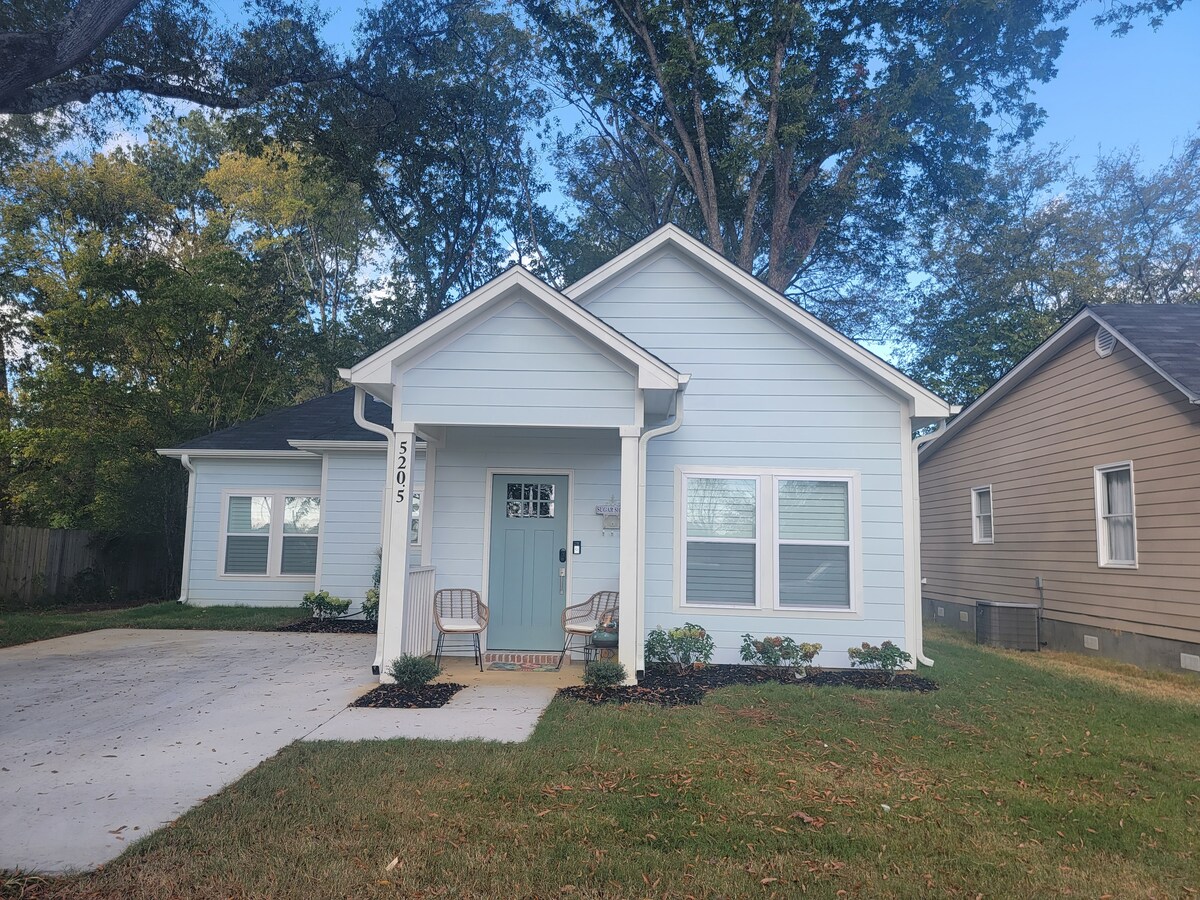 The exterior of the cottage displays soft blue siding with white trim and a welcoming front porch. Two comfortable chairs are positioned on the porch, inviting relaxation. A manicured lawn and landscaped flower beds enhance the charming curb appeal of the home.